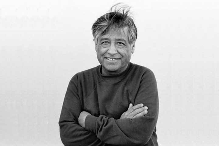 Black and white photograph of Cesar Chavez smiling with arms crossed wearing dark long-sleeved shirt.